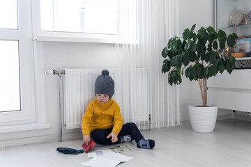 A little boy in a yellow sweater and hat is counting money and studying heating bills, near a heater with a thermostat..