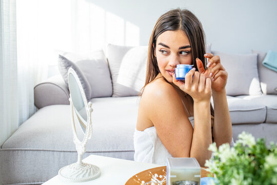 Woman applying face cream. Young happy woman applying moisturizer on her face. Skin care is the ultimate beauty. Young female in the bathroom looking in the mirror and taking care of her facial skin.