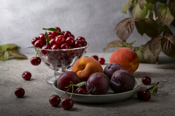 Cherry in a vase, peach and plum on a plate on a gray table.