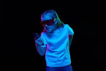Portrait of little beautiful girl, child in white T-shirt posing, shouting in phone isolated over black studio background in neon light