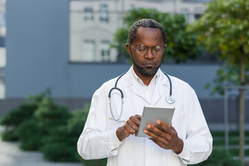Portrait of serious and focused doctor at work, african american man in glasses working with tablet computer outside clinic.