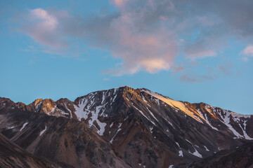 Scenic landscape with high snow mountain with sharp rocky pinnacle in golden sunlight under clouds of sunset color at changeable weather. Colorful view to large mountain top under cloudy sunset sky.