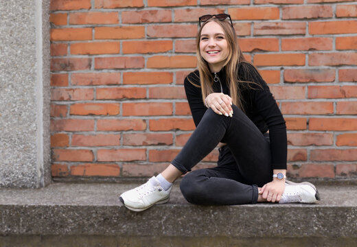 Portrait Of A Cheerful Cute Smiling Girl, 17 Years Old, With Long Hair And Sunglasses On Her Head, In A Black Turtleneck, Sits Against The Background Of A Brick Wall