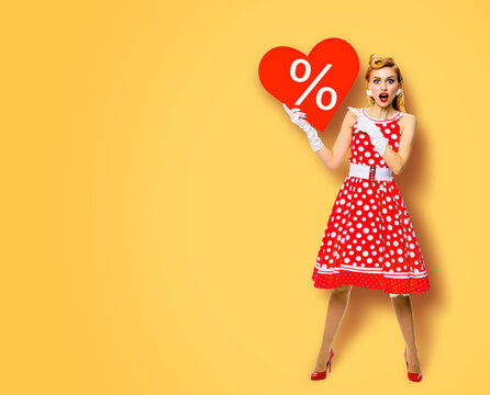 Woman holding red paper heart shape with % sign. Full length body portrait of happy pin up girl showig pointing signboard. Sales ad concept. Yellow background. Valentine or like symbol. Rockabilly.