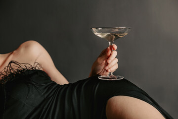Unrecognizable woman in evening black dress, lying on a black studio background and holding glass...