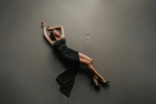 Above view of seductive woman, dressed in a slim elegant black dress with open shoulders and fashionable heels, lying on the floor near glass of drink in isolated dark studio