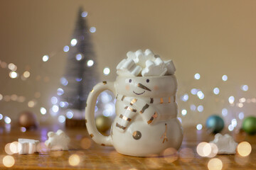 a snowman cup filled with white marshmallow snowflakes on a table against a background of a Christmas tree and holiday lights