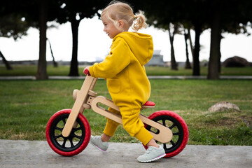 Happy active pretty cute caucasian blonde baby girl, kid, toddler,smiling child about 2 years old wearing bright yellow jumpers learning riding run balance bike in summer park outdoors.