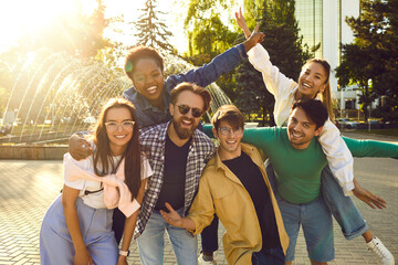 Group of good friends having fun together. Happy cheerful beautiful young diverse mixed race people standing on sunny city square, posing for photo, piggybacking girls, looking at camera and smiling