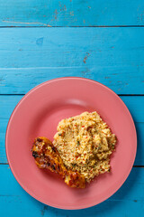 Couscous with vegetables in sauce and spices with spiced chicken drumstick on a pink plate on a blue table.copy space
