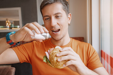 A man with a cheerful face pours an excess of salt on his burger in a restaurant. Risk of...