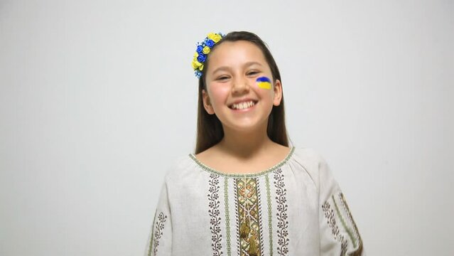 A girl in an embroidered dress and the flag of Ukraine on her face stands against the backdrop of a white wall and laughs. Girl in a good mood on a white background