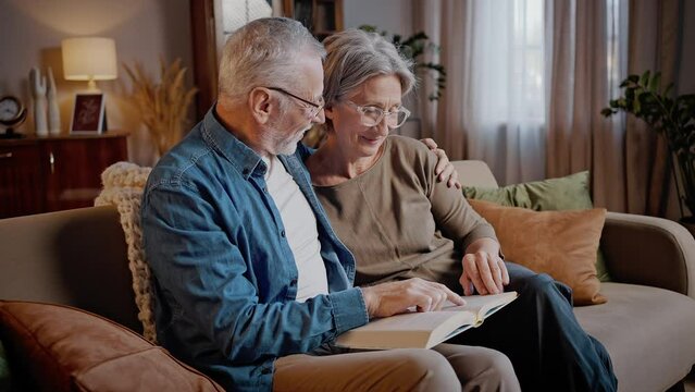 Happy elderly couple reads book together sitting on sofa