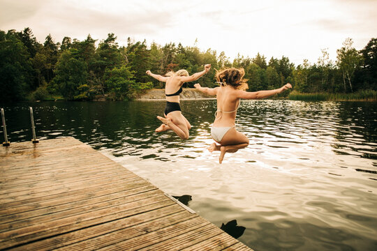 Female friends wearing bikini jumping in lake with arms outstretched during vacation