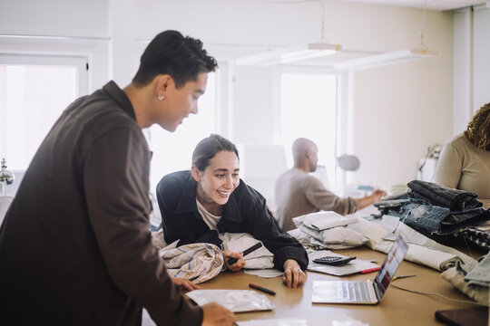 Smiling fashion designer looking at laptop by male colleague at workshop