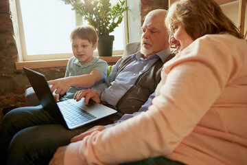 Life portrait of friendly family, grandparent and their grandson sitting on sofa and spending time together, using modern gadgets, talking, studying
