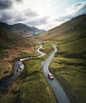 Red car travelling down a winding road next to a stream in the Lake District