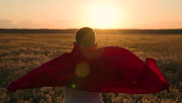 Son superhero in red cape in nature. Active Boy, child plays superhero in sun. Happy run of child in red raincoat at sunset in park. Kid runs across green field with flowers, childhood dream to fly.