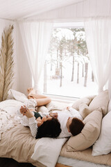 A happy couple in love in pajamas are resting, looking out the window at the forest and lying on the bed in a cozy Scandinavian-style bedroom in a country house in winter. Selective focus