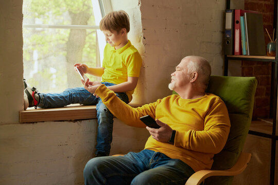 Grandfather and grandson sitting on chair and looking at digital tablet and cellphone. Relatives spending time together, teaching each other to use modern gadgets
