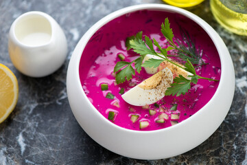 Cold beetroot soup with slices of boiled chicken egg and fresh greens served in a white bowl, horizontal shot, middle closeup