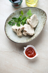 Bowl with georgian khinkali dumplings, fresh parsley and dipping sauce on a light-beige stone background, vertical shot, elevated view