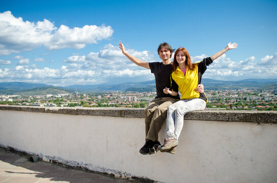 Happy tourist couple in love, young man and woman, sitting on tall building above city near mountains in Ukraine, laughing and raising their hands up, against background of blue sky and clouds