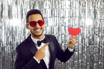 Attractive man in classic suit shows red heart in his hand standing on shiny silver background....