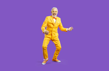 Happy funny old man in eccentric suit and glasses dancing on violet studio background. Smiling mature showman or performer in costumer laugh make dancer moves. Entertainment concept.