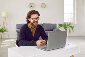 Positive brown hair Caucasian man owner of technology business with smile looks at laptop screen participating in meeting with company team or negotiations with partners sits at desk in home office