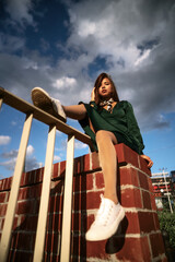 Street style fashion portrait of a cute young brown haired girl in a green dress under the cloudy sky. Attractive slim woman sits on the fence and poses in the light of the sun.