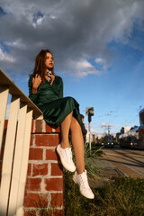 Street style fashion portrait of a cute young brown haired girl in a green dress under the cloudy sky. Attractive slim woman sits on the fence and poses in the light of the sun.
