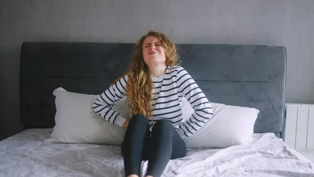 Caucasian woman suffering from severe stomach ache sitting on bed in bedroom after wake up feeling extremely sick. Young adult female experiencing period cramps. Girl suffering from sharp bladder pain