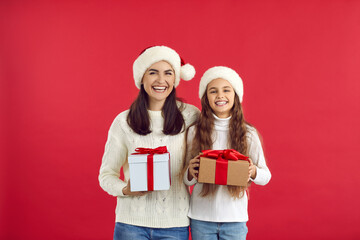 Portrait of smiling young Caucasian mother and teen daughter in Santa hats isolated on red studio background greet with New Year. Happy mom and girl child hold presents congratulate with Christmas.
