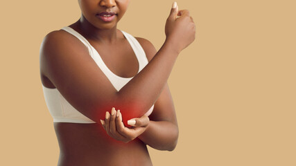 Black woman suffering from arthritis, stiff elbow, bruise, or tennis elbow condition holding hand on painful area on her arm isolated on beige color banner background. Injury, health problem concept