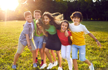 Group of happy cheerful active little children enjoying summer break, meeting in park, frolicking on green grass, playing fun games, making new friends, hugging, smiling and having great time together - obrazy, fototapety, plakaty