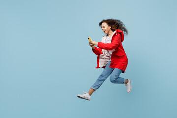 Obraz premium Full body side view young woman of African American ethnicity wear red jacket jump high run use mobile cell phone isolated on plain pastel light blue cyan background. Wet fall weather season concept.