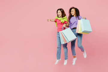 Full body young two friends women wear shirts hold in hand paper package bags after shopping jump high together isolated on pastel plain light pink color background. Black Friday sale buy day concept.