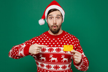 Merry shocked young man in red warm knitted sweater Santa hat posing hold in hand mock up of credit...