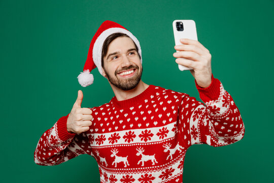 Merry young man wear red knitted sweater Santa hat posing doing selfie shot on mobile cell phone show thumb up isolated on plain dark green background studio New Year 2023 holiday celebration concept