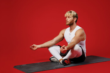 Plakat Full body young strong sporty sportsman man wear white clothes spend time in home gym sit on yoga mat meditate hold hands in om gesture isolated on plain red background Workout sport fit body concept