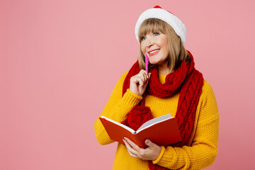 Merry minded elderly woman 50s year old wear yellow knitted sweater red scarf Santa hat posing hold book diary pen prop up chin isolated on plain pink background Happy New Year Christmas 2023 concept