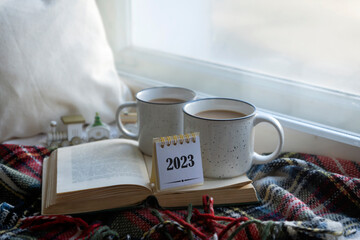 Calendar for 2023 and two mugs of fragrant hot coffee on the window with a blanket on a winter morning.