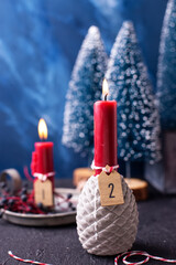 Christmas composition. Red burning advent candle with board with number two,  wild blue berries and blue decorative trees against blue  textured background. Scandinavian minimalistic style. 