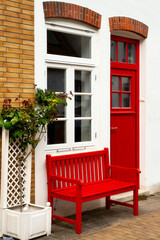 House entrance with red front door, white window ,under it a red bench and roses in flowerpot