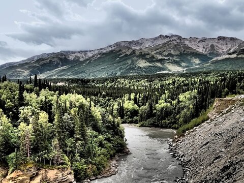 Alaska - River running through forest, with a rock
ledge on one side and mountains in the distance,
gloomy sky; photo manipulation for graphic effect