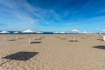 sunny sandy beach with numerous white wooden umbrellas against a blue cloudy sky