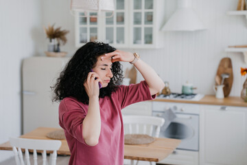 Young curly Italian woman talking by phone in lilac sweatshirt looking aside confidently. Caucasian pretty housewife at kitchen talking using smartphone. Serious arabic woman getting audio message.