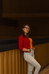 Young Latin woman with long hair dressed casually looks straight at the camera with a serious expression while leaning on a railing in a beautiful interior