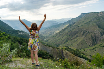 Fototapeta premium Young happy woman traveler in dress with outstretched arms standing on grassy hillside in summer mountains enjoying moment